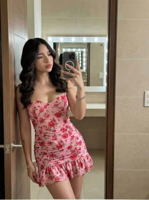 A girl in a pink rose tight dress takes a confident selfie in the mirror in the bathroom