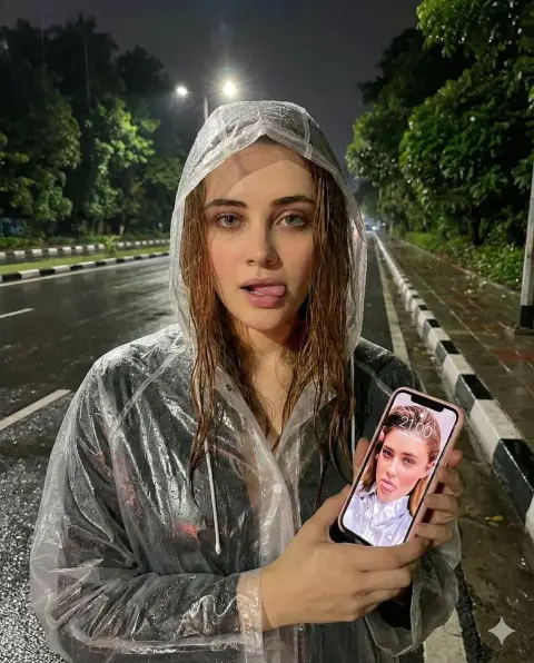 Close-up of playful girl in transparent raincoat holding mobile phone on the street on rainy night