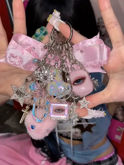 Use Image 1 as the reference for the keyring designMatch its design language, overall shape, silhouette, and layered charm composition. The keyring should look richly decorated and distinctly Y2K, with a pink glam kitsch mood. Include details like a heart charm, padlock, mini phone charm, plush charm, rhinestones, beads, glossy plastic pieces, acrylic charms, chrome metal hardware, and layered early-2000s accessory details.
Use Image 2 as the reference for the person. Match the girl’s styling, visual vibe, pose energy, and fashion mood.
Create a 7-second video.
Composition: the girl is placed behind the keyring. She raises both hands upward in front of her head, with her hands close enough to create a visible open space or “frame” between them. The keyring is positioned inside that hand-framed space, in front of her, as if suspended there or gently held between her hands. Do not place the keyring flat over the center of her face. Her face should remain partially readable behind it, with the keyring occupying the framed zone between her hands.
Motion sequence: at the beginning, the keyring is centered in the foreground inside the space created by her raised hands. She gently rotates or tilts the keyring, making the charms shimmer and swing slightly. Then, near the end of the video, she slowly moves the keyring sideways or slightly downward, revealing her full face clearly behind it. After the reveal, she looks toward the camera with a soft, stylish expression while her hands still frame the keyring nearby.
The keyring should remain the main foreground focal point for most of the video, but the final moment should clearly show the girl’s face. The interaction should feel delicate, intentional, and editorial, as if she is presenting a precious Y2K accessory.
Overall mood: Y2K cute gyaru, pink glam kitsch, glossy, playful, decorative, fashion-editorial, early-2000s accessory mood. Emphasize shiny charm movement, glossy plastic reflections, rhinestone sparkle, chrome hardware, pink glam styling, and a polished nostalgic girly aesthetic.
Camera: close-up portrait framing, slight slow push-in, shallow depth of field, keyring sharp in the foreground, girl visible behind it. Smooth motion, no sudden cuts.