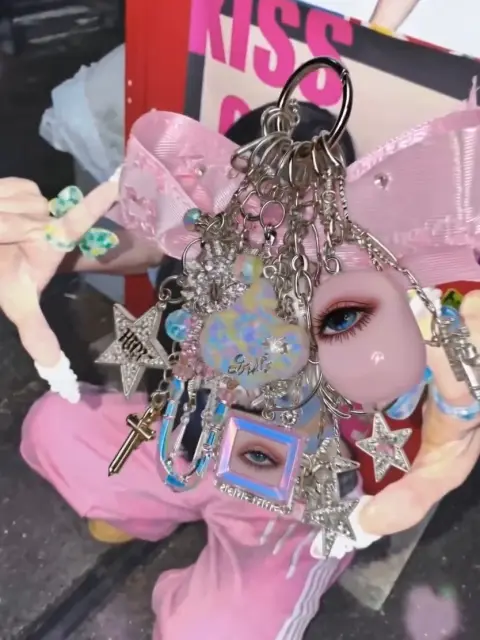 Use Image 1 as the reference for the keyring designMatch its design language, overall shape, silhouette, and layered charm composition. The keyring should look richly decorated and distinctly Y2K, with a pink glam kitsch mood. Include details like a heart charm, padlock, mini phone charm, plush charm, rhinestones, beads, glossy plastic pieces, acrylic charms, chrome metal hardware, and layered early-2000s accessory details.
Use Image 2 as the reference for the person. Match the girl’s styling, visual vibe, pose energy, fashion mood, and overall composition. Retain the original composition of Image 2 as much as possible, rather than replacing it with a completely different pose.
Create a 7-second video.
Composition: the girl is placed behind the keyring, and the keyring hovers in the foreground within the gesture space created by her hands. Do not make her physically hold the keyring. Preserve the original pose structure from Image 2. If the figure in Image 2 has one hand posed near her face, keep that composition and make the hand read as a soft palm-up presenting gesture, as if supporting the hovering keyring space or zone. If the figure in Image 2 has two hands posed together or framing the face, place the keyring hovering inside the open zone created by those hands. In all cases, do not place the keyring flat over the center of her face. Her face should remain partially readable behind it, with the keyring floating slightly in front of her.
Motion sequence: at the beginning, the keyring hovers in the foreground in the presentation zone near her face and hands. It gently rotates or tilts, making the charms shimmer and swing slightly. The girl remains poised and stylish behind it. Near the end of the video, the keyring slowly drifts sideways or slightly downward, revealing her full face clearly. After the reveal, she looks toward the camera with a soft, stylish expression while the keyring remains nearby in the frame.
The keyring should remain the main foreground focal point for most of the video, but the final moment should clearly show the girl’s face. The interaction should feel delicate, intentional, and editorial, as if she is presenting a precious Y2K accessory without directly gripping it.
Overall mood: Y2K cute gyaru, pink glam kitsch, glossy, playful, decorative, fashion-editorial, early-2000s accessory mood. Emphasize shiny charm movement, glossy plastic reflections, rhinestone sparkle, chrome hardware, pink glam styling, and a polished nostalgic girly aesthetic.
Camera: close-up portrait framing, slight slow push-in, shallow depth of field, keyring sharp in the foreground, girl visible behind it. Smooth motion, no sudden cuts.